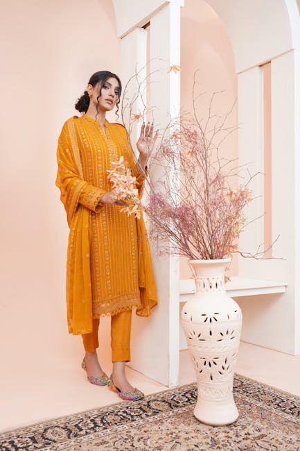 Stylish young woman modeling traditional Pakistani clothing indoors by vase with dried flowers.